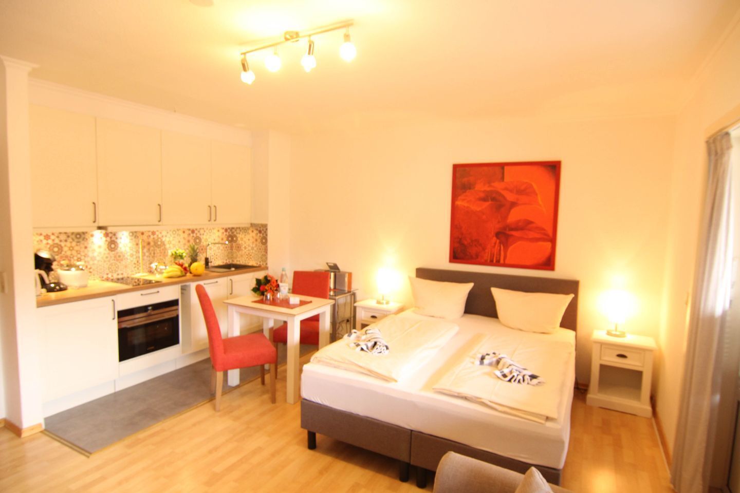 A hotel room with a bed, bedside tables, a desk with a chair, a kitchenette, and a red-framed painting on the wall.