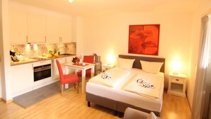 Une chambre d’hôtel comprenant un lit, des tables de chevet, un bureau avec une chaise, une kitchenette et un tableau encadré de rouge accroché au mur.