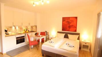 A hotel room with a bed, bedside tables, a desk with a chair, a kitchenette, and a red-framed painting on the wall.