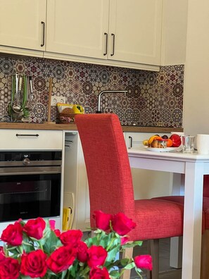 A kitchen with white cabinets, a patterned backsplash, a red chair, and a clock.
