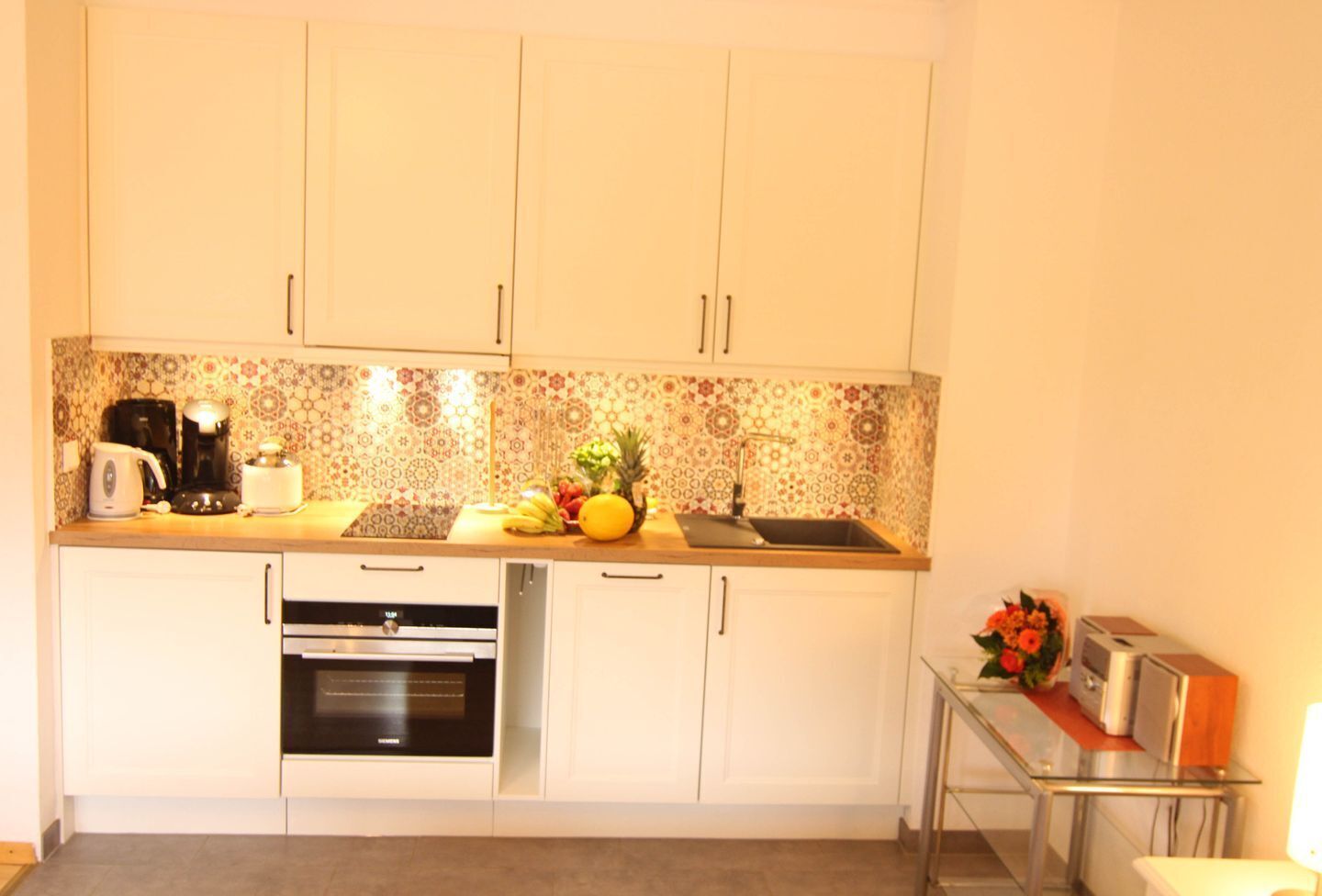 Een compacte keuken met witte kasten, een decoratieve spatwand, een ingebouwde oven, een vaatwasser en een klein aanrecht met een koffiezetapparaat, fruit en een broodrooster.