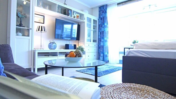 Ein modernes Wohnzimmer mit einer Couch, einem Couchtisch mit Obstschale, einem Flachbildfernseher und einem Bücherregal.