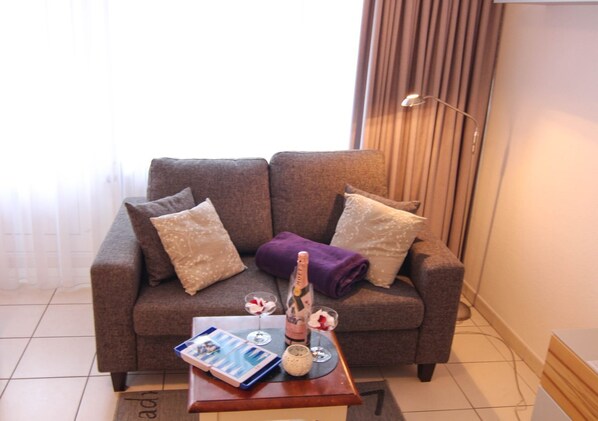A modern living room with a grey sofa, two decorative pillows, a purple throw blanket, a small table with a book, a glass of wine, and a candle.