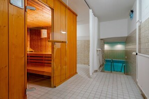 Et sauna rum med trævægge og en glasdør, der fører til et fliset badeværelse med et bruseområde med blå fliser.