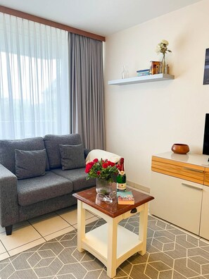 Una sala de estar con un sofá gris, una pequeña mesa de centro de madera con un jarrón de flores rojas, un televisor sobre un mueble, y una ventana con cortinas.