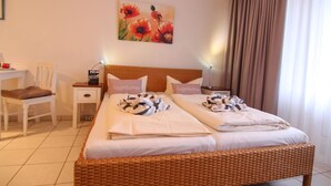 Ein Hotelzimmer mit einem Doppelbett, zwei Nachttischen, einem Sessel und einem Blumenbild, sowie einem Fenster mit Vorhängen.