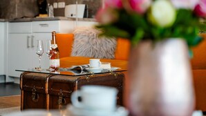 Un espacio de sala acogedor con una maleta antigua sobre una mesa de vidrio, una botella de vino y una taza. Un mullido cojín naranja y un jarrón con flores complementan la decoración.