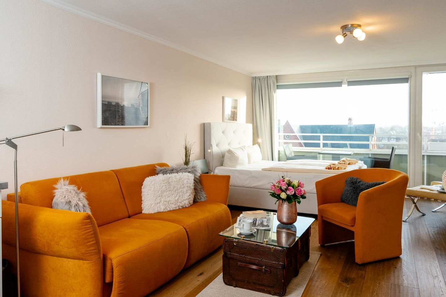 Un salon moderne avec un canapé orange, un fauteuil assorti, une table basse en verre et un lit donnant sur un paysage urbain.