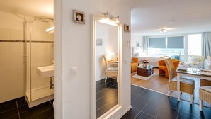 Habitación de hotel con ducha, un espejo grande, zona de comedor con mesa y sillas, y vista al mar.