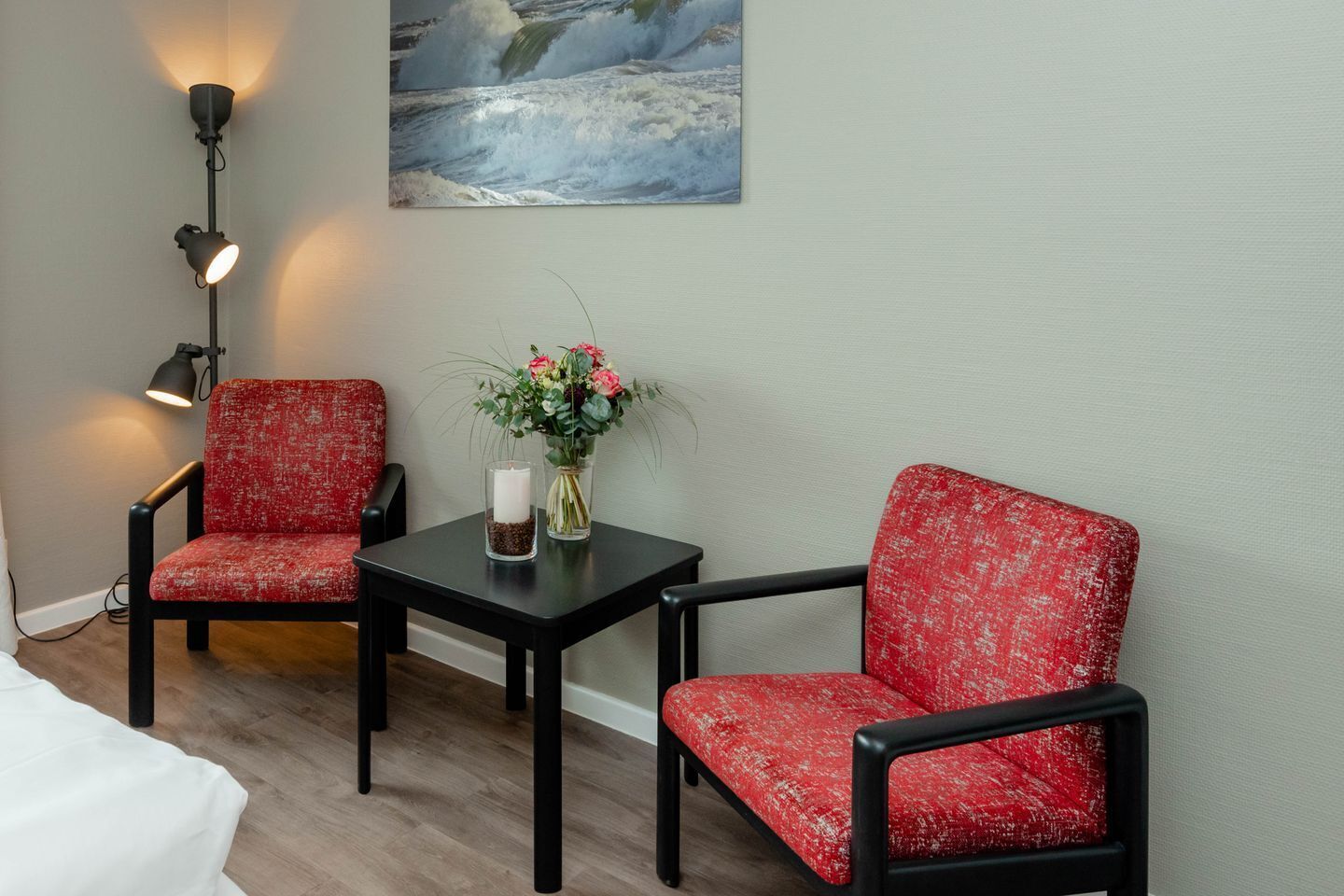 Ein Zimmer mit zwei roten Polstersesseln, einem kleinen schwarzen Tisch mit Kerze und Blumen, einem Gemälde einer Welle und einer Stehlampe.