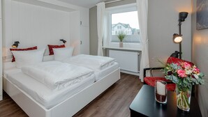 Una cama doble con ropa de cama blanca, una mesita con un florero y una ventana con cortinas.