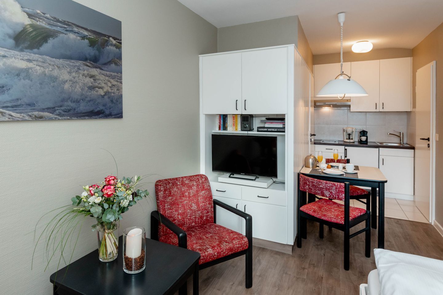 Een compacte hotelkamer met een televisie, een klein zitehoekje, een kitchenette en een schilderij van een golf aan de muur.