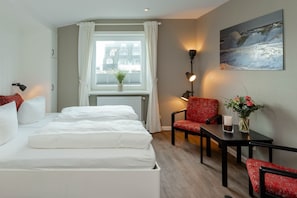 Une chambre d’hôtel avec un lit, deux chaises, une table avec un vase, une lampe et une fenêtre avec des rideaux.