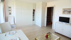 Un soggiorno moderno con un divano bianco, un tavolino da caffè in vetro con frutta, una televisione a schermo piatto su un mobile bianco e un'ampia armadio a muro con scaffalature integrate.