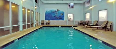 Una piscina interior con agua azul cristalina, un mural de delfines y varias sillas de descanso a lo largo de la piscina.