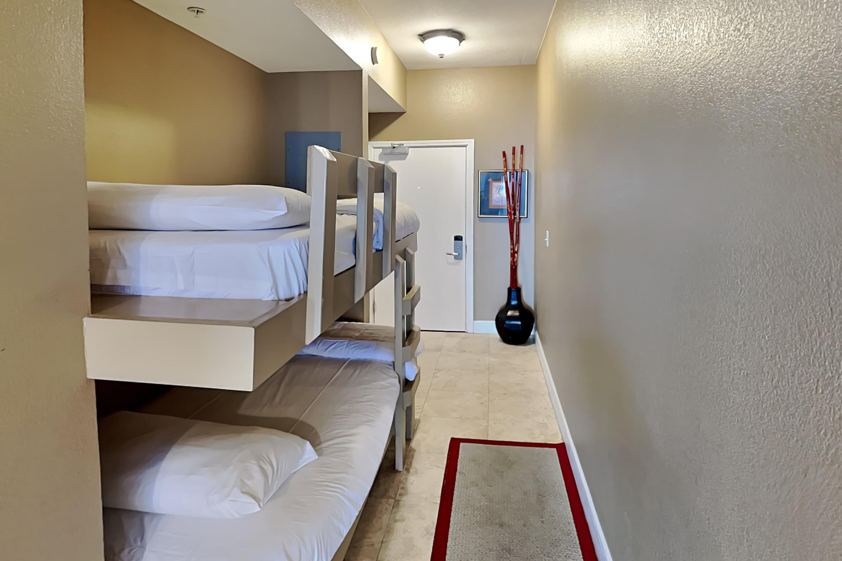 Una habitación de hotel con literas, una puerta, un cuadro colgado en la pared y una alfombra roja y blanca.