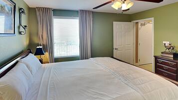 A bedroom with a bed, a ceiling fan, a window with curtains, a door, and a dresser with a lamp.