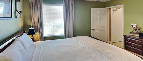 A bedroom with a bed, a ceiling fan, a window with curtains, a door, and a dresser with a lamp.