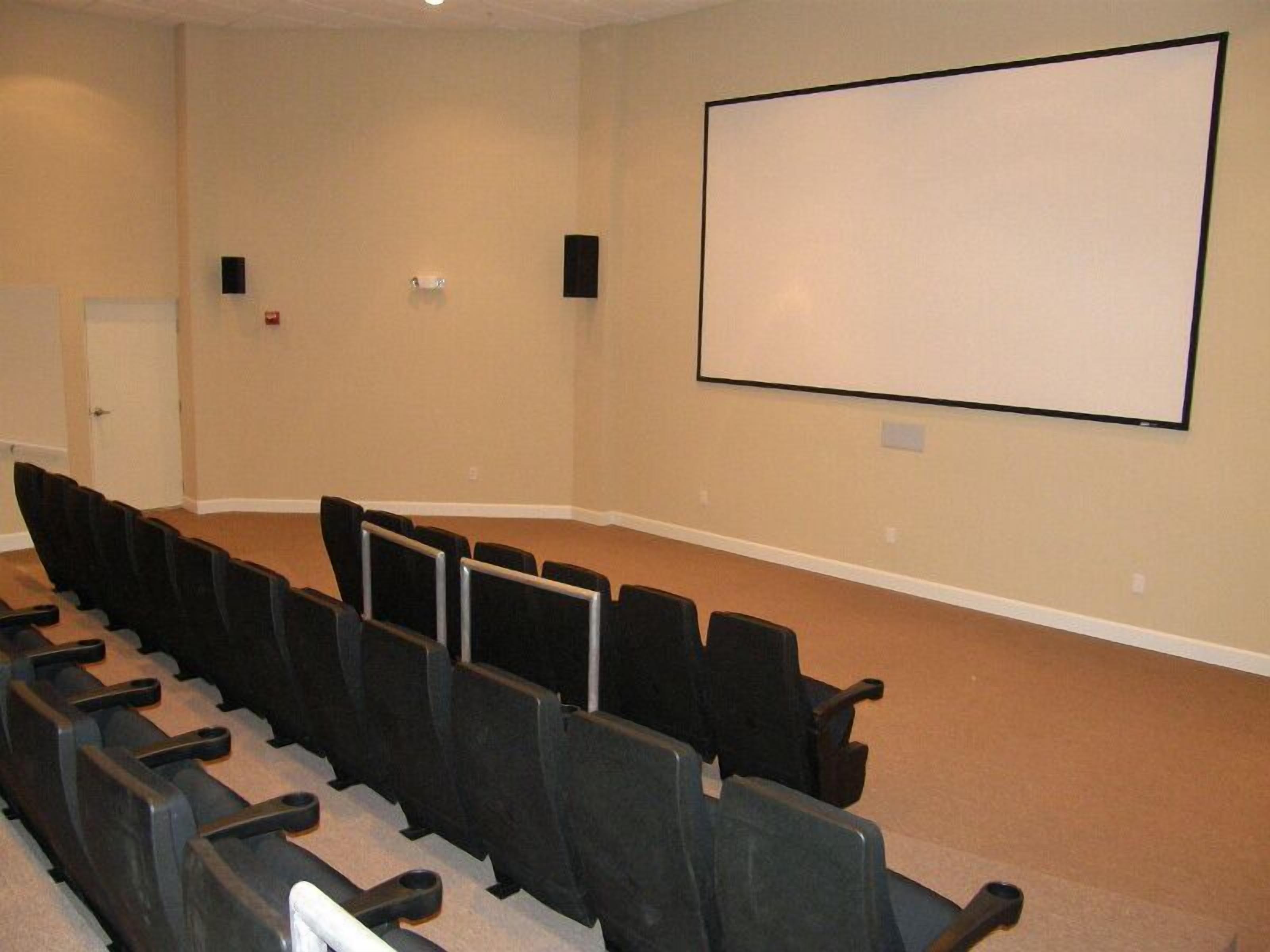 A conference room with rows of black chairs, a whiteboard, and two speakers on the walls.