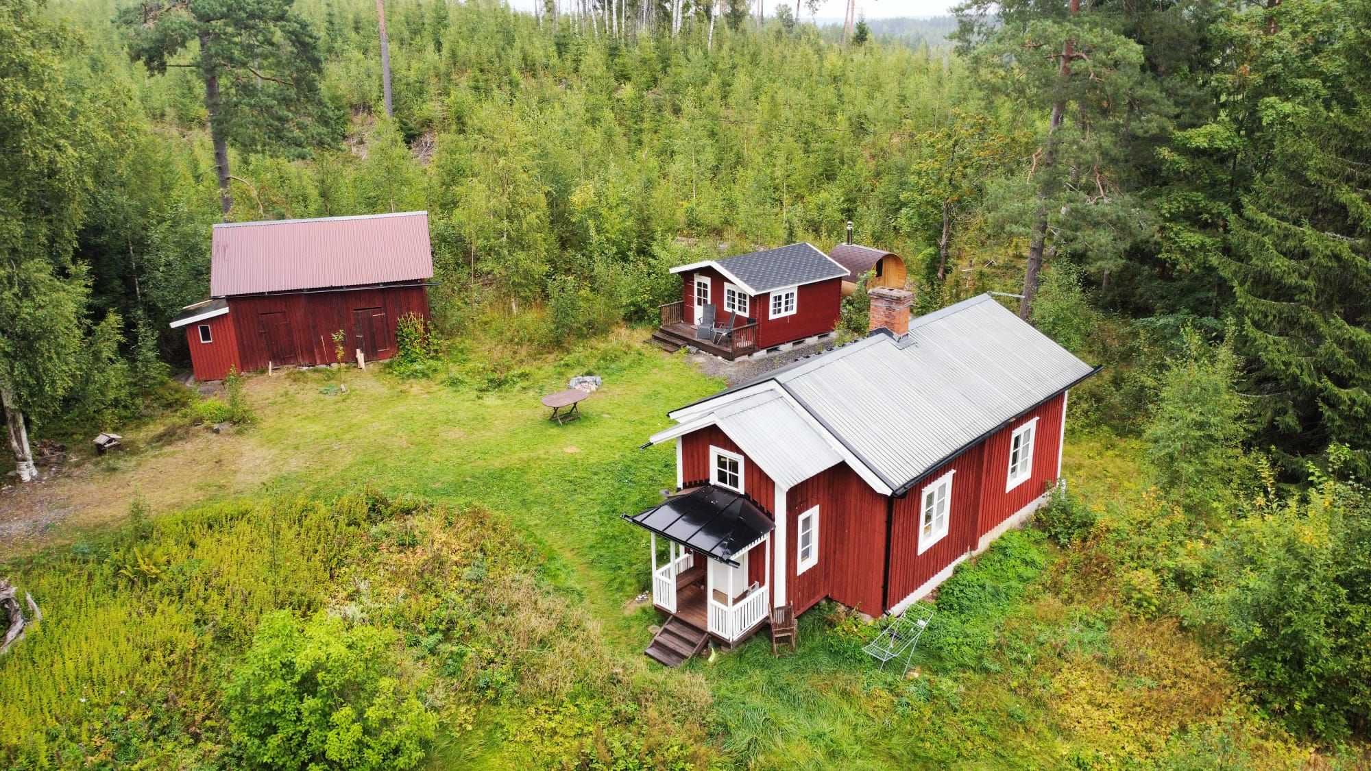 Ett rött trähus med en veranda och ett separat förråd som ligger i ett skogsområde.