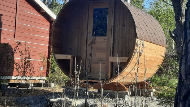 Een houten, vatvormige hut met een deur en raam, omgeven door bomen en een rood gebouw.
