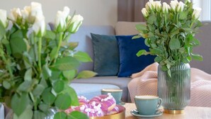 A cozy living room with a sofa, a coffee table with cups and a tray of snacks, and two vases with flowers.