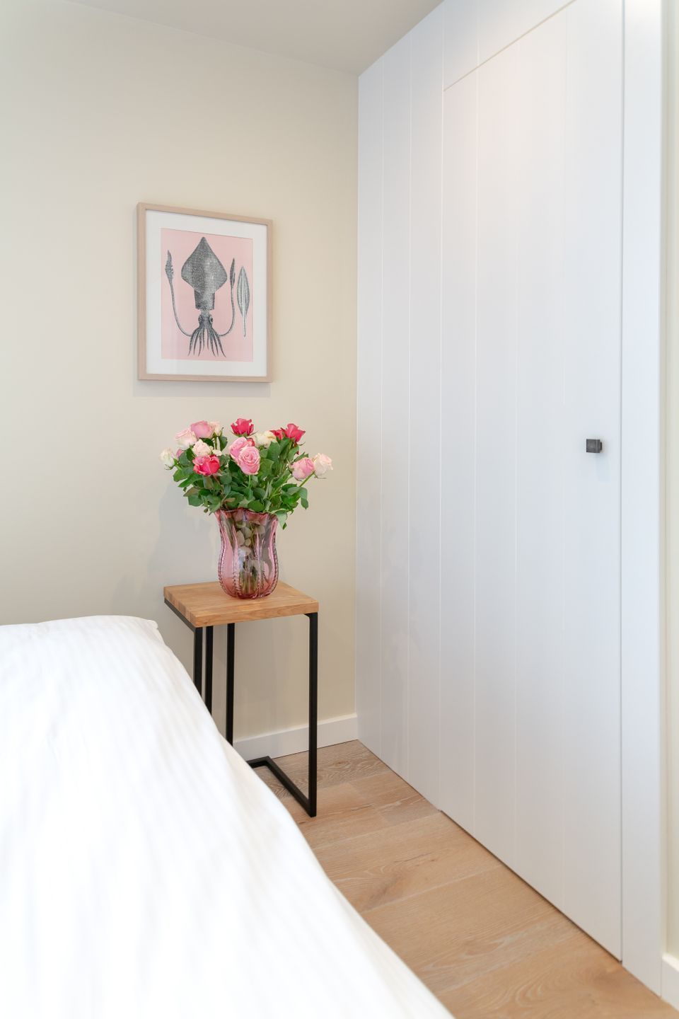 Ett sovrum med en säng, ett litet träbord med en vas med blommor, en vit dörr och en inramad tavla på väggen.