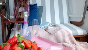 A cozy room with a striped curtain, a bed with a blue pillow, a pink tray with strawberries, a vase with pink roses, and a bottle.