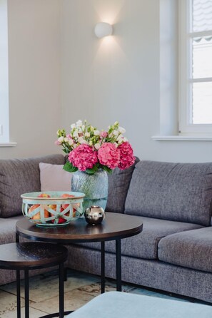 A living room with a grey sofa, a round coffee table, a vase with pink flowers, and a fruit basket.