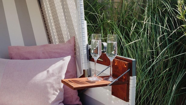 Een gezellig buiten zitgedeelte met een comfortabele bank, een houten tafel met twee glaasjes en een fles, en uitzicht op hoge grassen en bloemen.