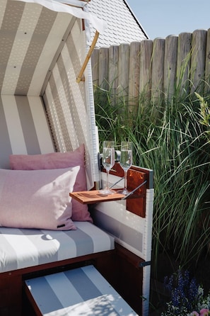 A cozy outdoor seating area with a cushioned bench, a wooden table with two glasses and a bottle, and a view of tall grasses and flowers.