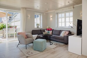 A modern living room with a grey sofa, a light blue ottoman, and a coffee table. There is a TV on a stand to the right and a window with white shutters.