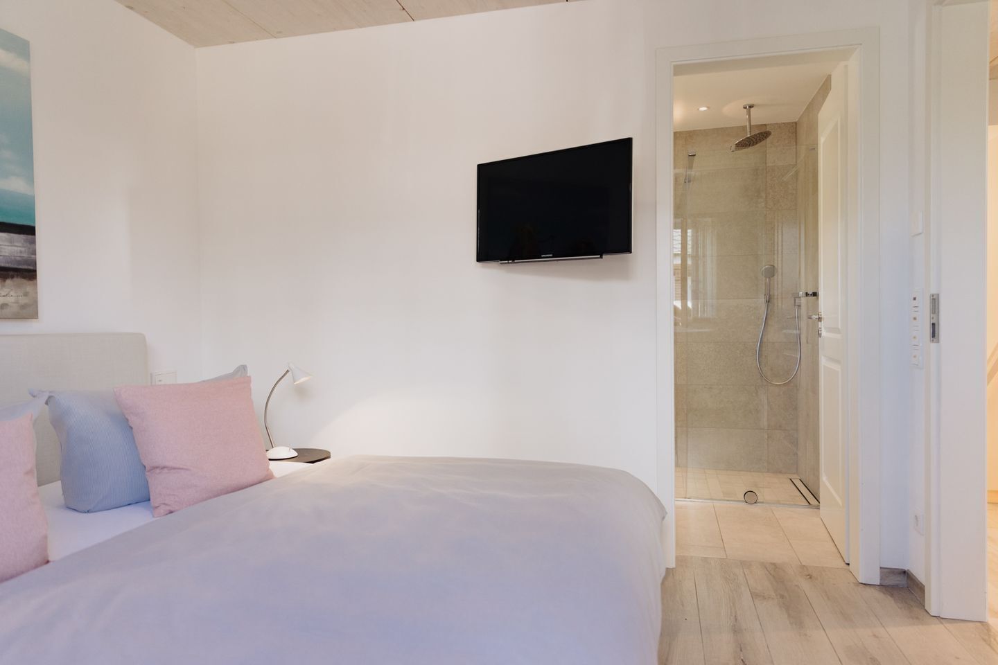 A bedroom with a bed, a TV mounted on the wall, and a shower area visible through an open door.