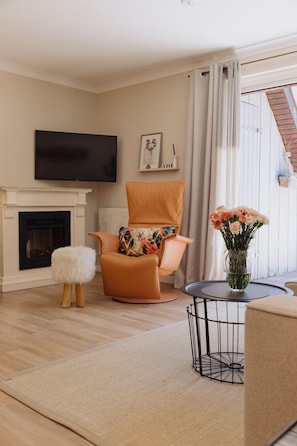 Un salón con chimenea, un televisor montado en la pared, un sillón naranja cómodo, una mesita redonda con un jarrón de flores y vistas al exterior a través de un ventanal grande.