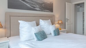 A neatly made bed with white and teal pillows, a large abstract painting above it, and a bedside table with a lamp and a glass of orange juice.