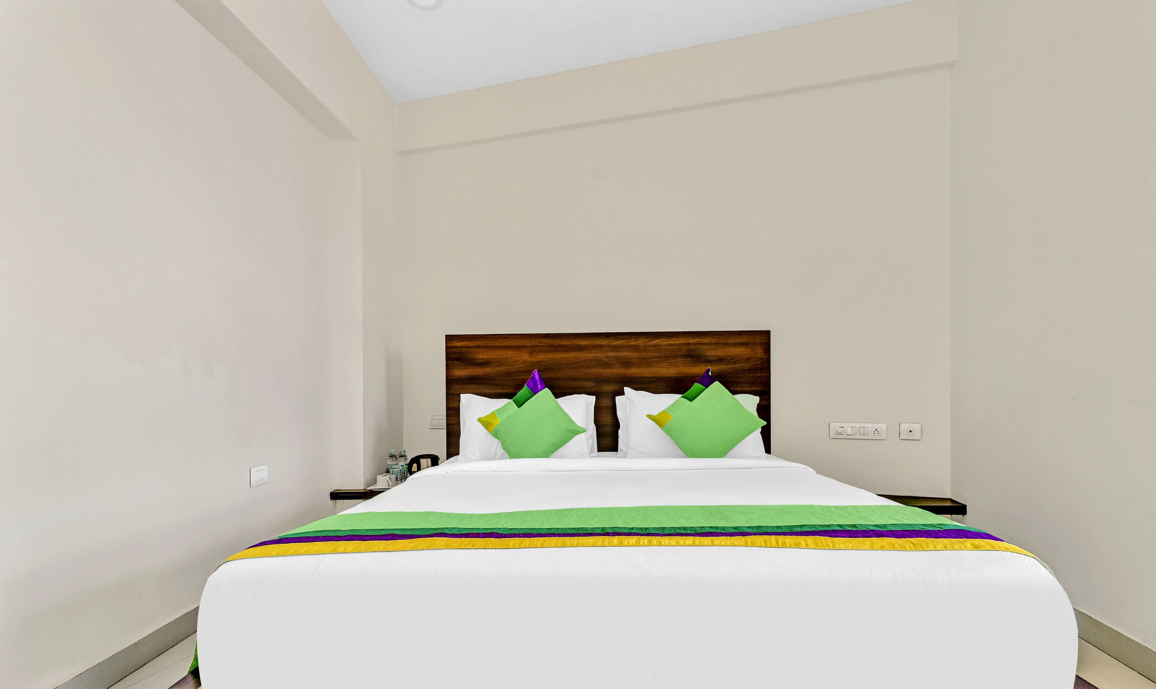 Een hotelkamer met een houten hoofdeinde, een bed met wit beddengoed, groene en gele kussens, en een nachtkastje met een lamp.