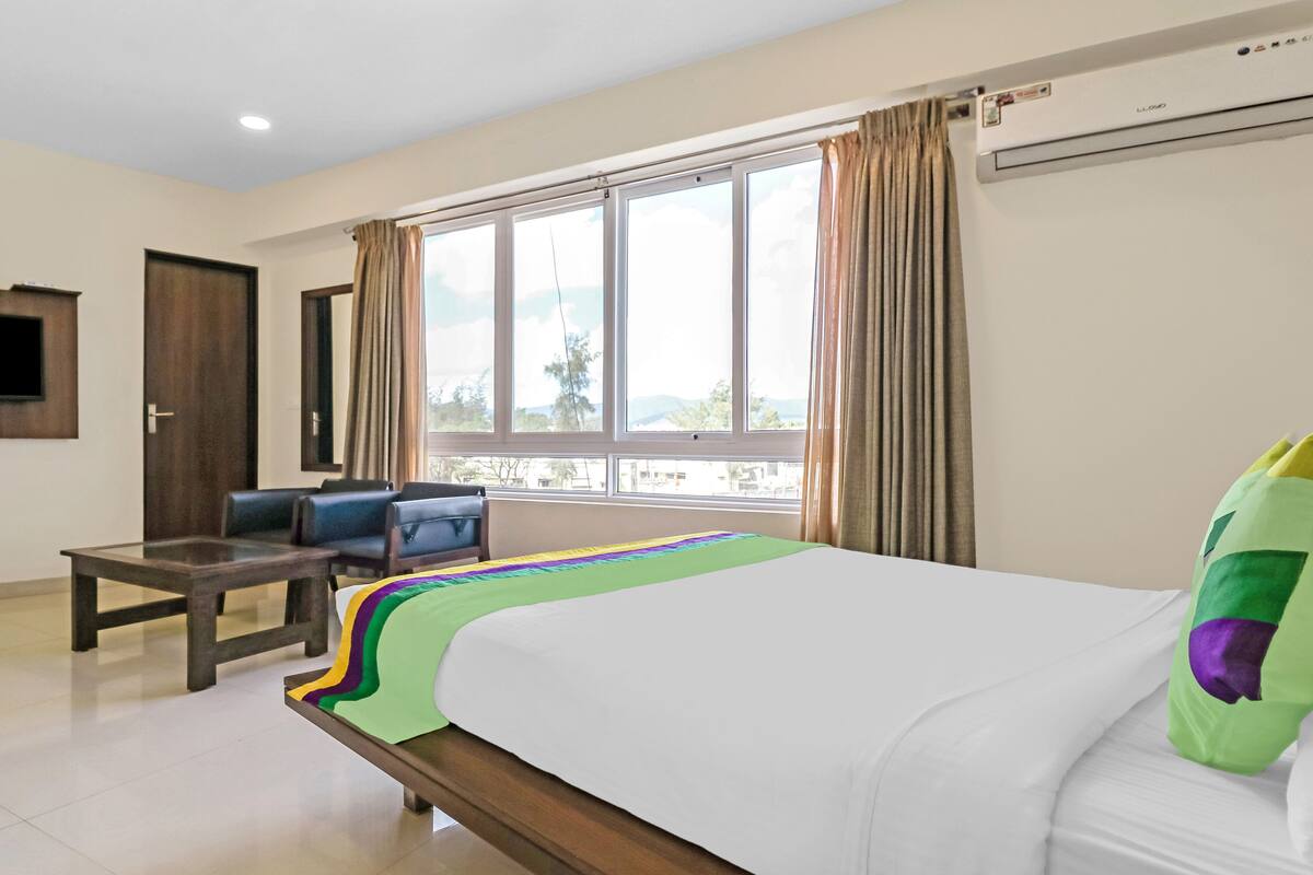 Quarto de hotel com cama, escrivaninha, cadeira, televisão e janela com cortinas.
