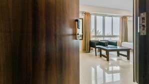 A hotel room with a wooden door, a dining area with a table and chairs, a large window with curtains, and a white wall with a clock. - Treebo Punarnava Bypass Road (Chikkamagaluru)