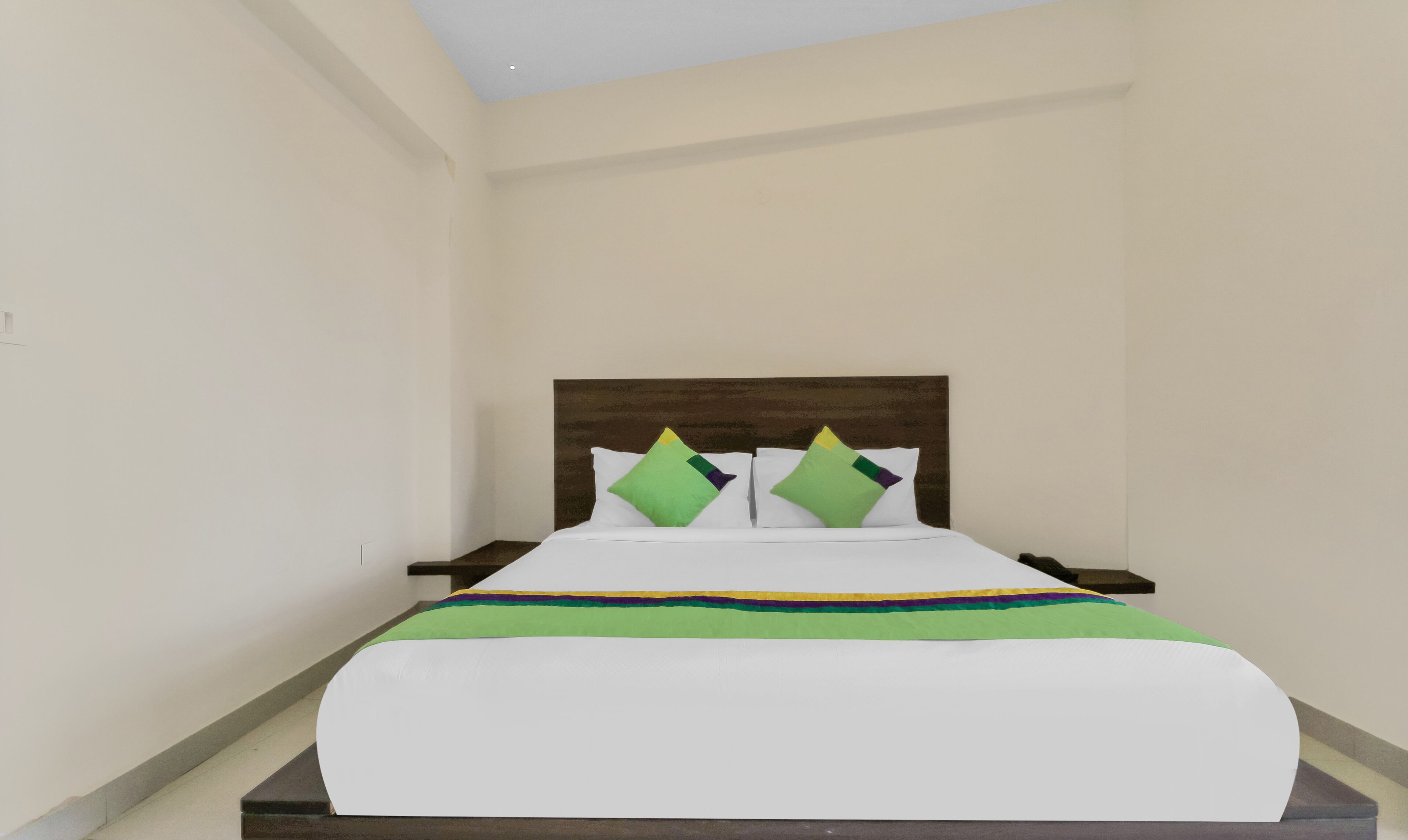 A neatly made bed with a white cover and a colorful striped blanket, two green pillows, and a wooden headboard.