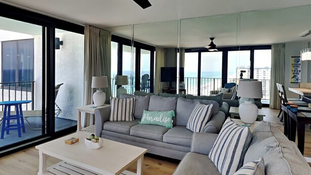 Un salon moderne avec un canapé gris, des coussins à rayures et une table basse blanche. Il y a un balcon avec une chaise bleue et une table.