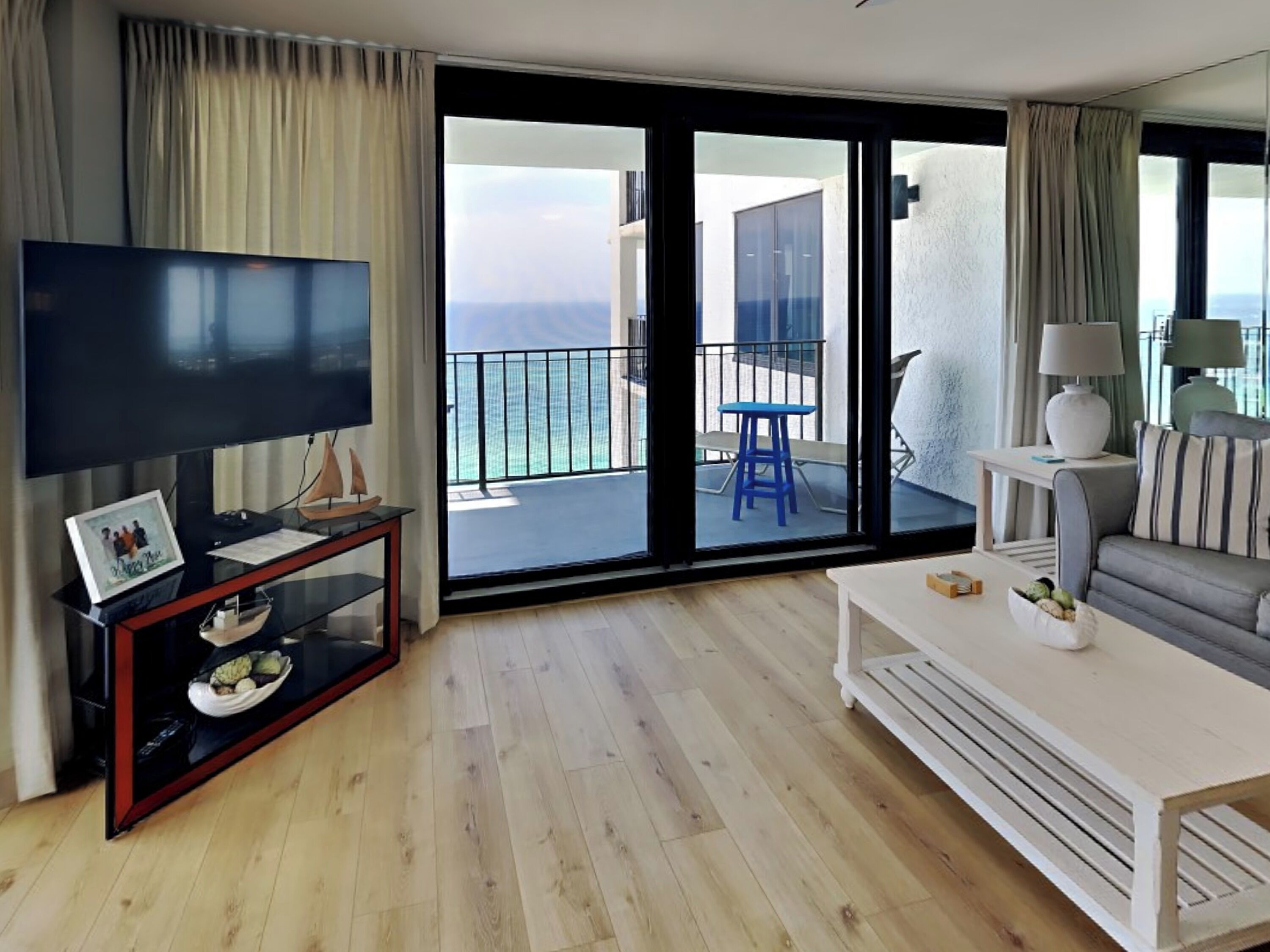 A modern living room with a large flat-screen TV, a white sofa, and a balcony with a view of the sea.
