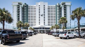 Un grand hôtel avec une entrée principale, de nombreux étages et un parking contenant plusieurs véhicules.