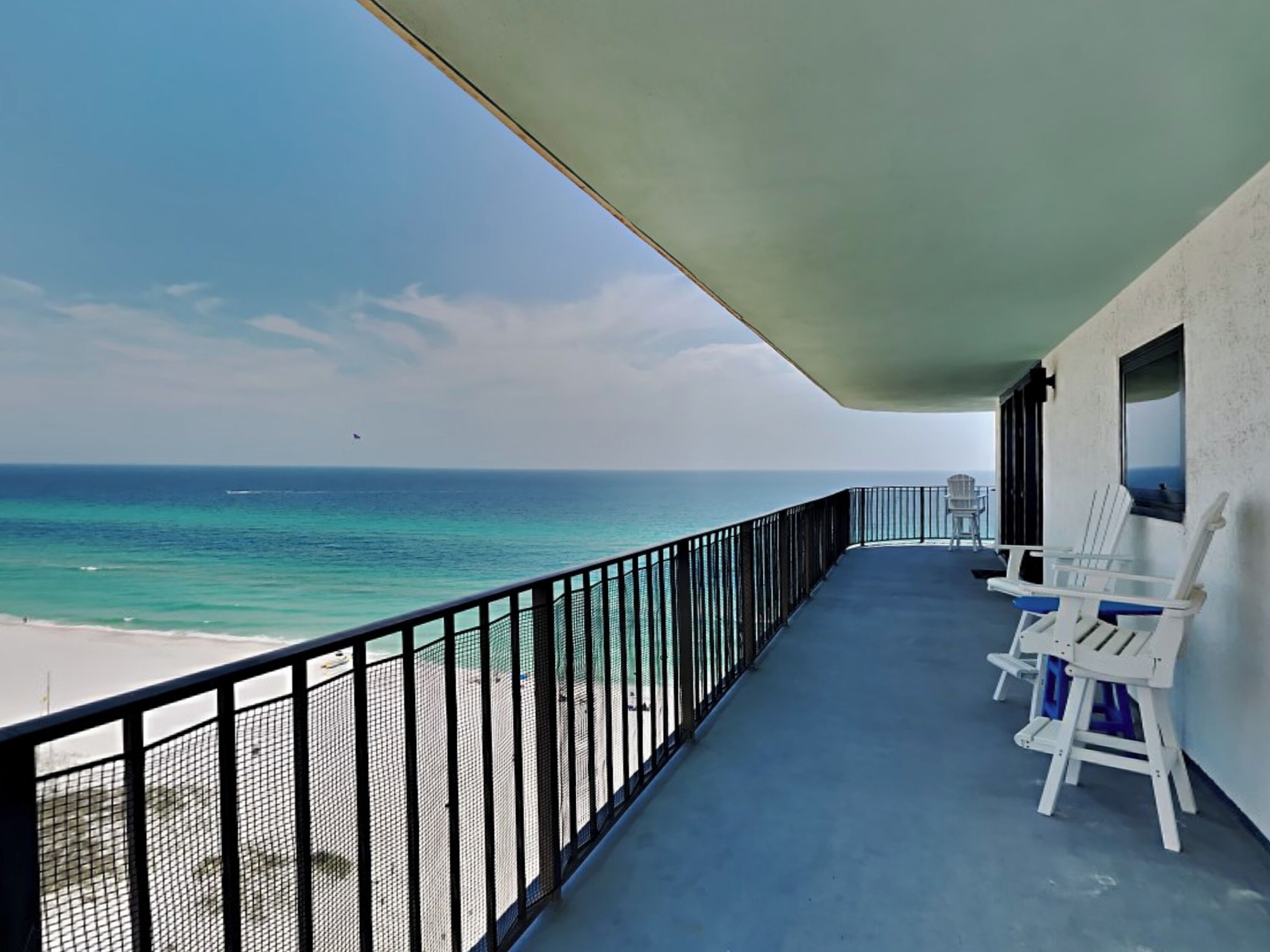 A balcony with a view of the ocean and beach. There are two white chairs and a table on the balcony.