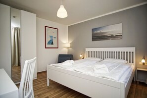 Une chambre d’hôtel avec un lit blanc, un bureau, une chaise, une lampe et un tableau au mur.