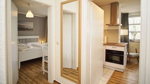 A bedroom with a bed, bedside table, and a hanging light fixture. There is a door leading to a bathroom with a mirror and a toilet. The room has wooden flooring and white walls.