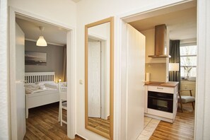 A bedroom with a bed, bedside table, and a hanging light fixture. There is a door leading to a bathroom with a mirror and a toilet. The room has wooden flooring and white walls.