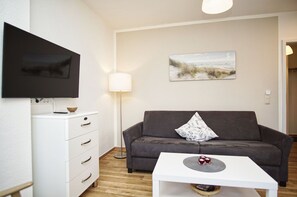 A modern living room with a grey sofa, a white coffee table with red candles, a flat-screen TV mounted on the wall, a floor lamp, and a painting of a beach scene.