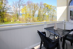 A balcony with a table and chairs, white railing, and a view of trees and a body of water. - Close to the beach, with balcony - Villa Strandmuschel FeWo 07 - Villa Strandmuschel (Göhren)