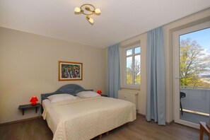 Un dormitorio con cama, mesita de noche, lámpara roja, un cuadro, ventana con cortinas y vista a árboles.