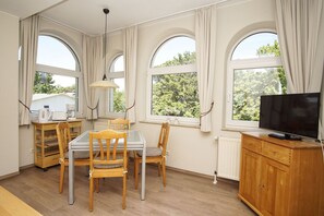 Una habitación con una mesa de comedor, sillas, televisor y ventanas grandes con cortinas.